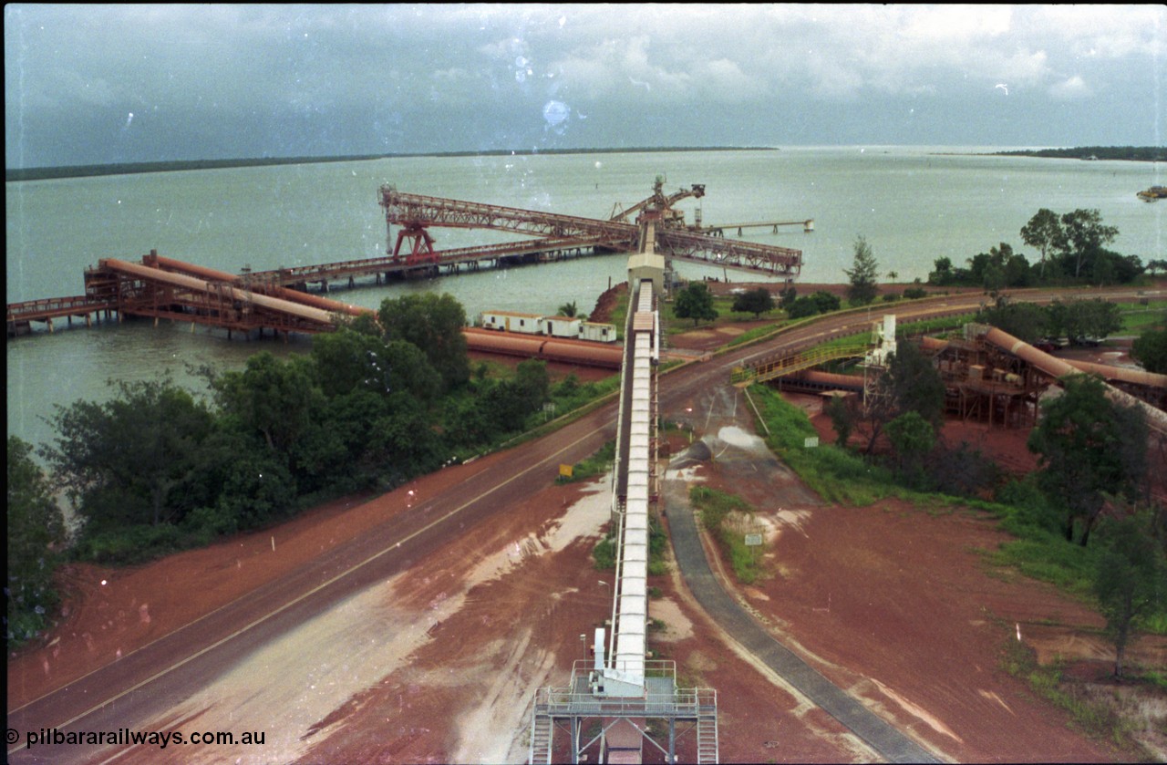 211-00
Weipa, Lorim Point, view from the top of the kaolin storage silos looking north at the kaolin conveyor and ship loader, with red legs and the No.2 bauxite loader straight ahead in the cyclone tie-down position. The bauxite conveyors can be seen passing under the road.

