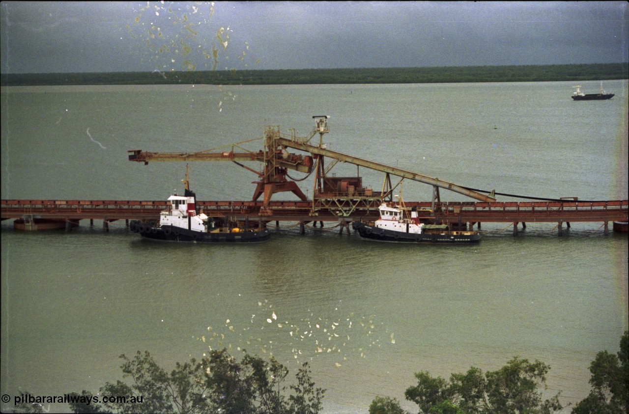 211-01
Weipa, Lorim Point, view from the top of the kaolin storage silos looking west at No.1 bauxite ship loader and the two tug boats. The unloading barge can be seen in the background in the Embley River.
