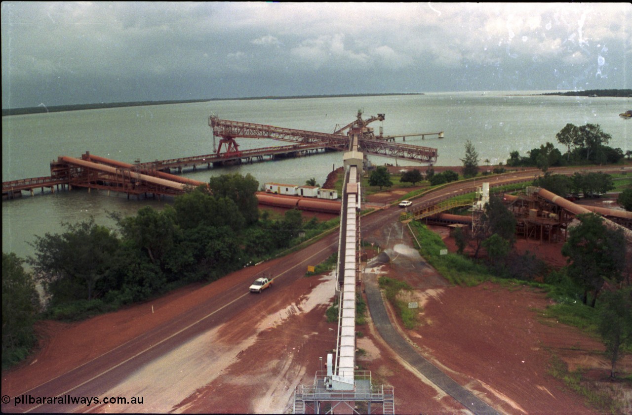 211-0A
Weipa, Lorim Point, view from the top of the kaolin storage silos tanks looking north at the kaolin conveyor and ship loader, with red legs and the No.2 bauxite loader straight ahead in the cyclone tie-down position. The bauxite conveyors can be seen passing under the road.
