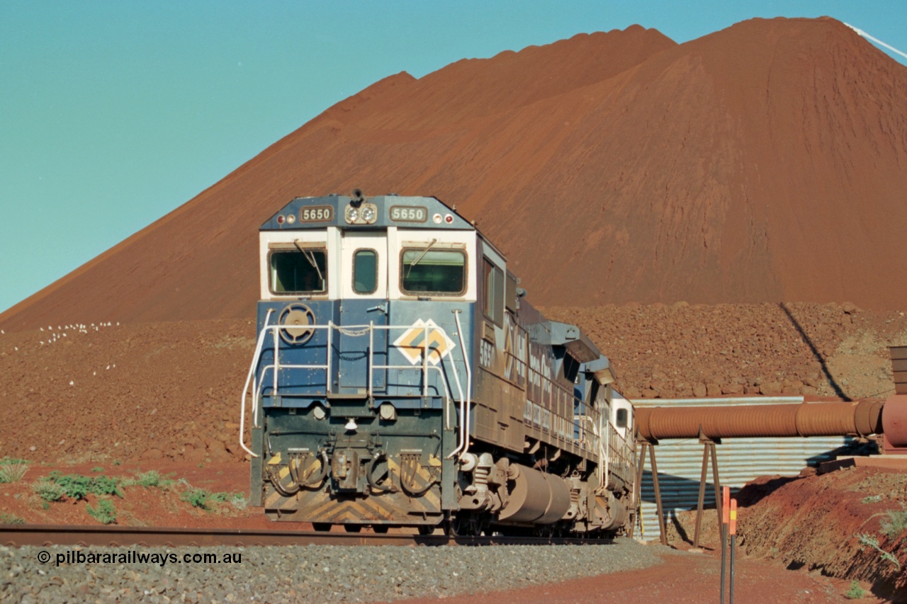 229-21
Yandi Two loadout, looking at the exit portal of the tunnel as Goninan rebuild CM40-8M GE unit 5650 'Yawata' serial 8412-07 / 93-141 drags a loading train through at 1.2 km/h, the massive pile of ore is gravity fed into the waggons via two sets of hydraulic chutes, the original pedestal radial stacker is visible above the ore along with the extraction fan. [url=https://goo.gl/maps/jmtnauf76Zq]GeoData[/url].
Keywords: 5650;Goninan;GE;CM40-8M;8412-07/93-141;rebuild;AE-Goodwin;ALCo;M636C;5481;G6061-2;