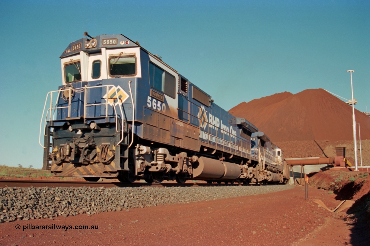 229-22
Yandi Two loadout, looking at the exit portal of the tunnel as Goninan rebuild CM40-8M GE unit 5650 'Yawata' serial 8412-07 / 93-141 drags a loading train through at 1.2 km/h, the massive pile of ore is gravity fed into the waggons via two sets of hydraulic chutes, the original pedestal radial stacker is visible above the ore along with the extraction fan. [url=https://goo.gl/maps/jmtnauf76Zq]GeoData[/url].
Keywords: 5650;Goninan;GE;CM40-8M;8412-07/93-141;rebuild;AE-Goodwin;ALCo;M636C;5481;G6061-2;