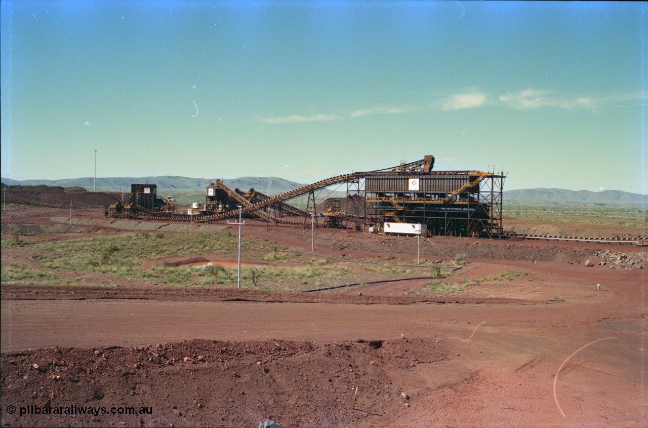 232-00
Yandi Two crushing and screening plant, built and operated by Henry Walker for BHP Iron Ore, overview from the radial stacker feed conveyor. From the left is the primary crusher, the dual secondary crushers can be made out behind the tertiary crushers in the middle and the screenhouse is the large building tot he right, the conveyor leading to the right is CV210. February 1997. [url=https://goo.gl/maps/TUReAkezgfz]GeoData[/url].
