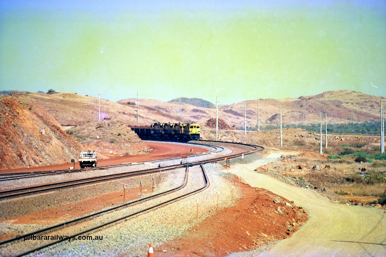 246-07
Cape Lambert, yard view of the then new extension to the south for the West Angelas mine was coming on stream as a Robe River loaded Deepdale train arrives on the main behind the standard quad Dash 8 power with 202 waggons, the just visible siding (rejoining) on the far left will become a compressor waggon holding road, the 6 km and Boat Beach Rd grade crossing are around the corner to the left. Closest to the camera is the extended No. 1 Road which becomes the Empty Car Line from car dumper one for the Deepdale traffic. Next is the original line or Mainline and the third line across is the Loaded Car Line for the West Angelas traffic to the new car dumper 2. 22nd May 2002.
