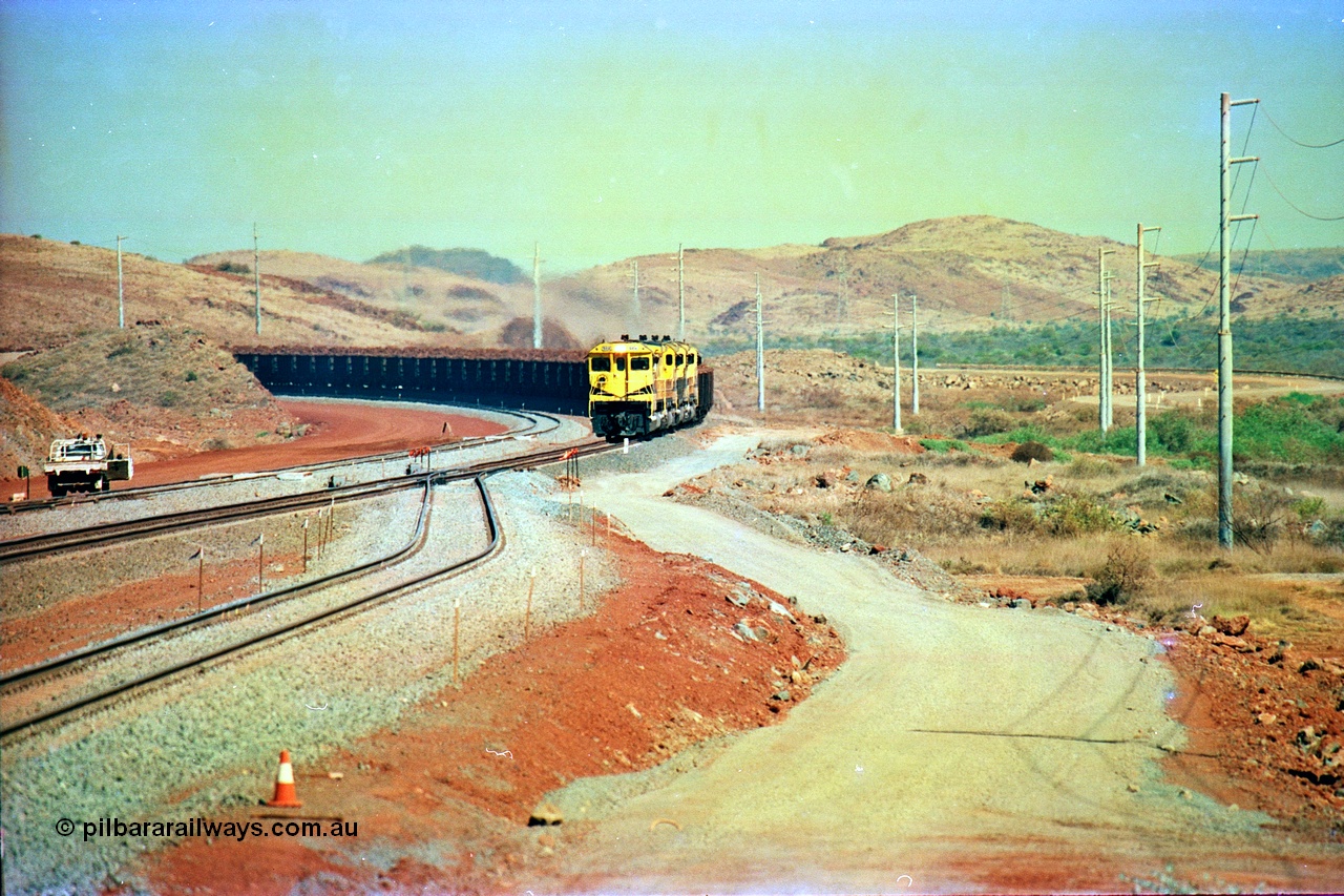 246-10
Cape Lambert, yard view of the then new extension to the south for the West Angelas mine was coming on stream as a Robe River loaded Deepdale train arrives on the main behind the standard quad Dash 8 power with 202 waggons, the just visible siding (rejoining) on the far left will become a compressor waggon holding road, the 6 km and Boat Beach Rd grade crossing are around the corner to the left. Closest to the camera is the extended No. 1 Road which becomes the Empty Car Line from car dumper one for the Deepdale traffic. Next is the original line or Mainline and the third line across is the Loaded Car Line for the West Angelas traffic to the new car dumper 2. 22nd May 2002.
