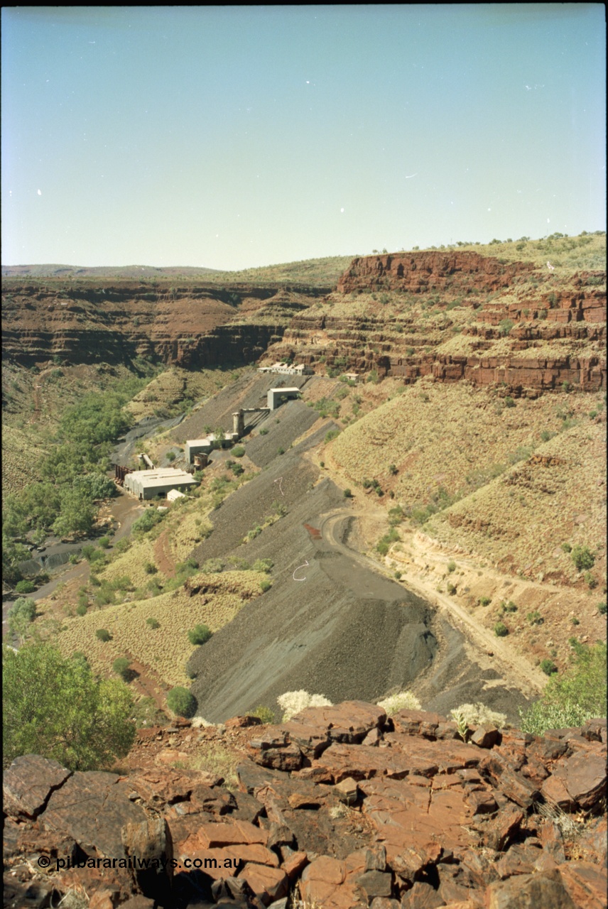 195-22
Wittenoom, view from above the Devil's Staircase of Colonial Mill and Mine with piles of tailings, milling site overview, looking south, railway workshops and underground offices are the higher buildings, then the primary crushing shed, holding silo, down to the mill and then the bag house and associated sheds, tailings line the gorge side.
