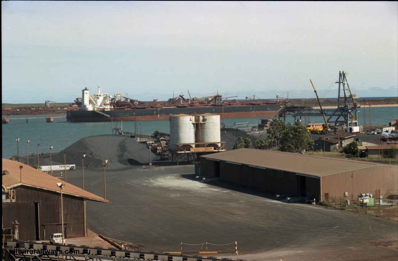198-28
Port Hedland Port, view of manganese stockpile area with truck unloading, Cockburn Cement silos, old loader can just be made out in front of new bulk loader under construction, 2001.
