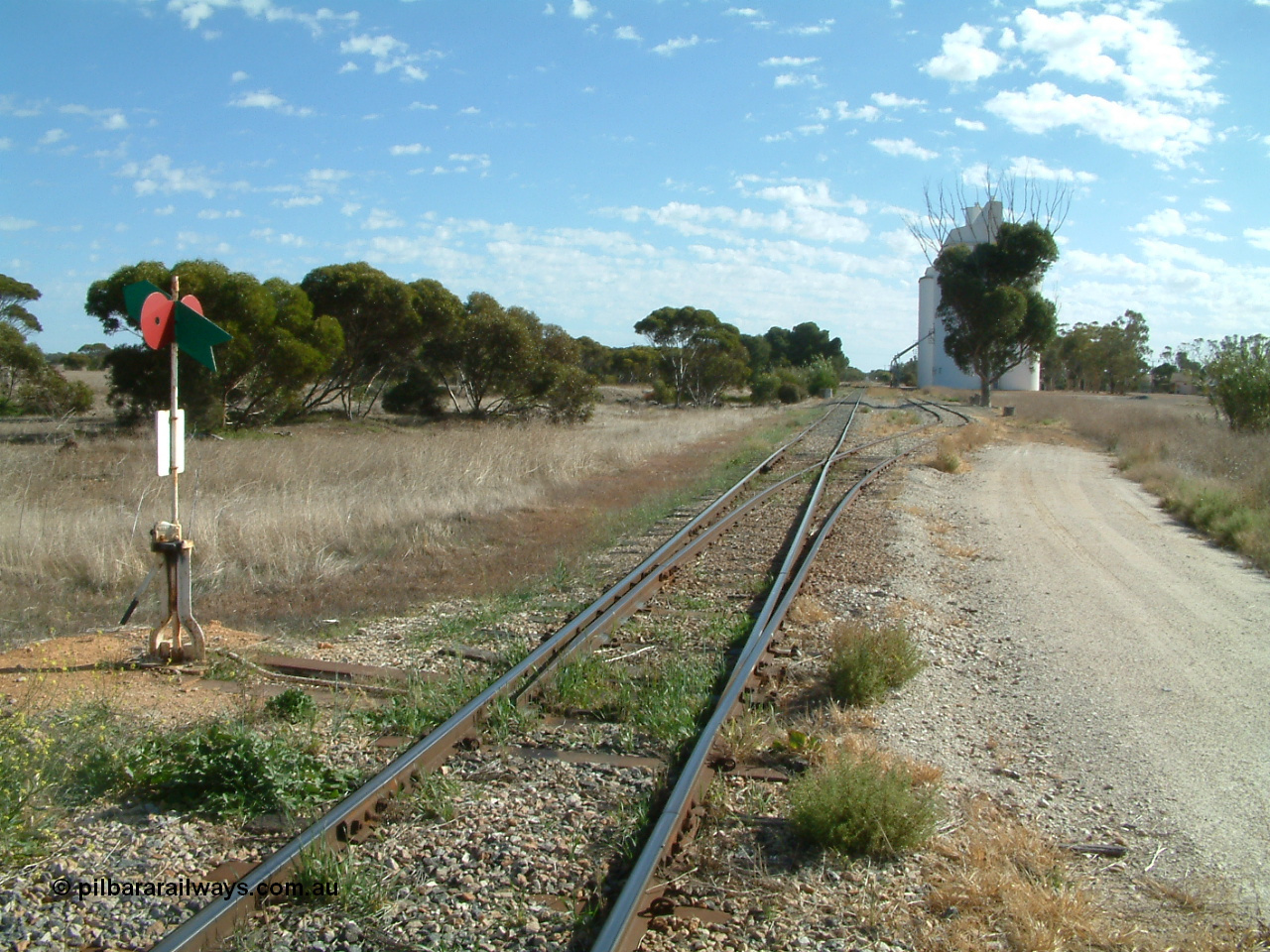 030406 093000
Yeelanna, station located at the 81.7 km, original terminus of line in April 1909, then junction for line to Mount Hope in October 1914. Station yard overview from the south end looking north, points, lever and indicator for the grain siding with concrete silo complexes in the background. The branch line to Kapinnie and former Mount Hope is just visible through the trees on the raised embankment. 6th April 2003.
