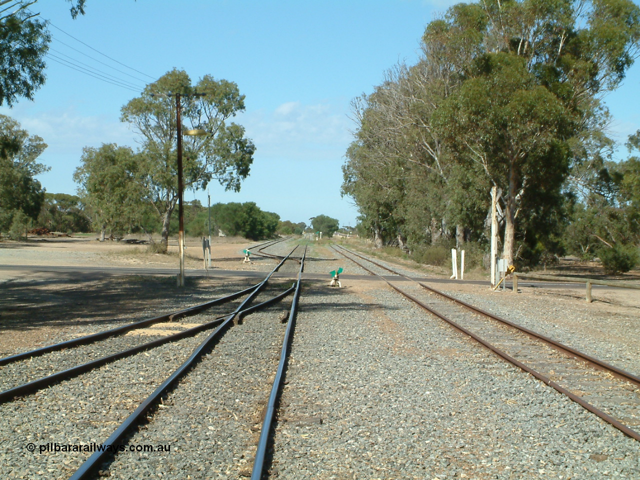 030406 100006
Cummins, yard view looking south across Railway Tce grade crossing, Buckleboo line - siding converging on the left, with the grain siding on the right, the points, indicator and lever south of the grade crossing is to the grain bunker loading - unloading site which is behind the trees to the left. 6th April 2003.

