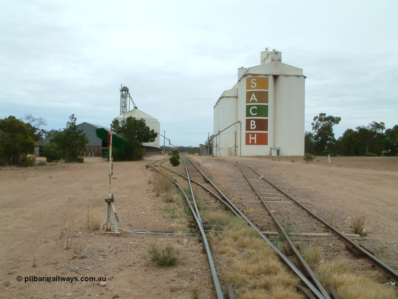 030407 104705
Poochera, station yard overview looking north from south end of Goods Siding 2, point lever and indicator for siding, on the left is the horizontal grain shed with an Ascom style silo complex behind it, on the right the goods shed can be made out along with the SACBH concrete silo complexes. 7th April 2003.
