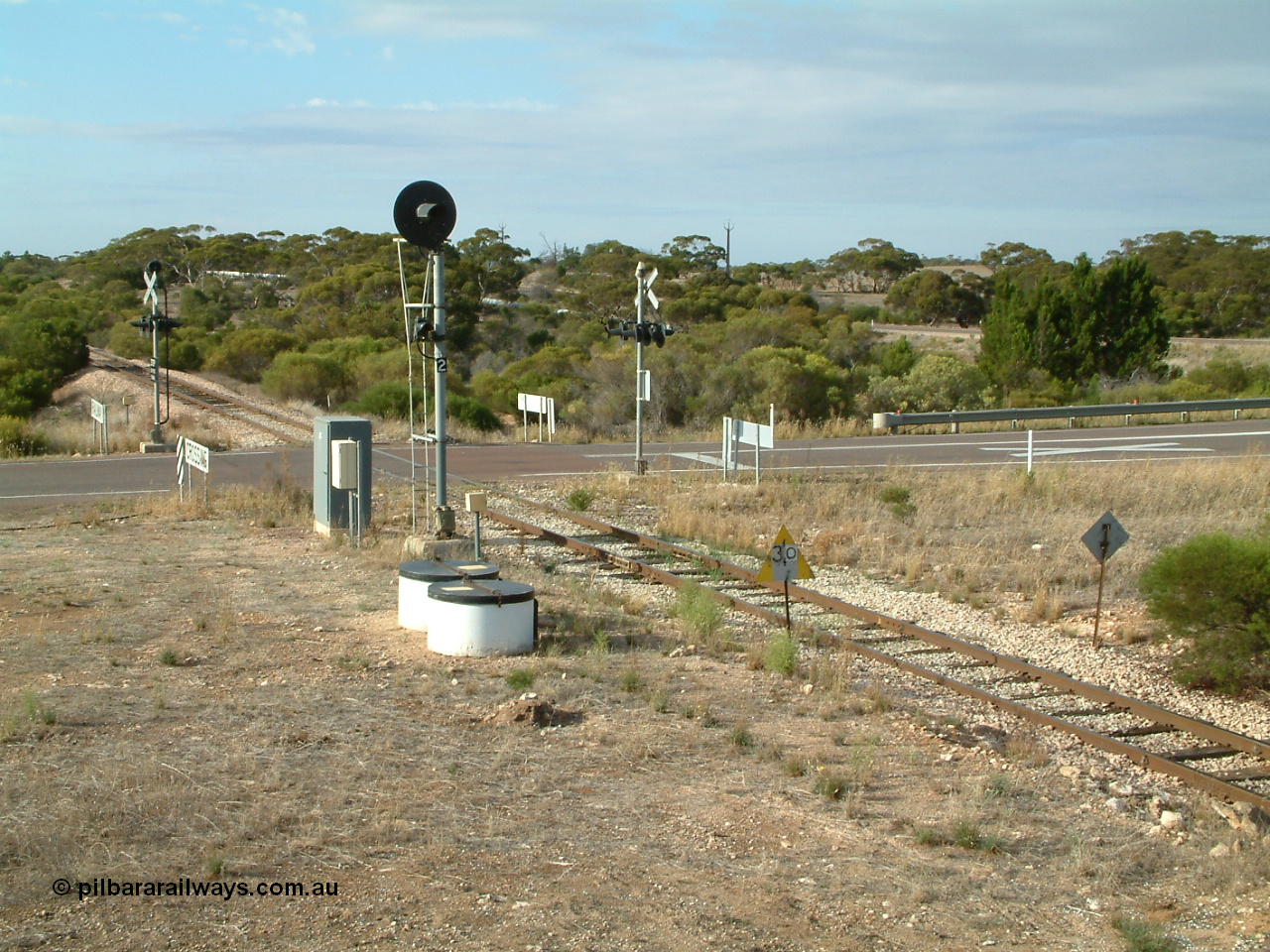 030409 083147
Kyancutta, view of the Eyre Highway grade crossing protected by 'F' type flashing lights, the searchlight signal 2 is one of only three on the Eyre Peninsula system, it is to allow Up trains to enter and shunt the yard.
