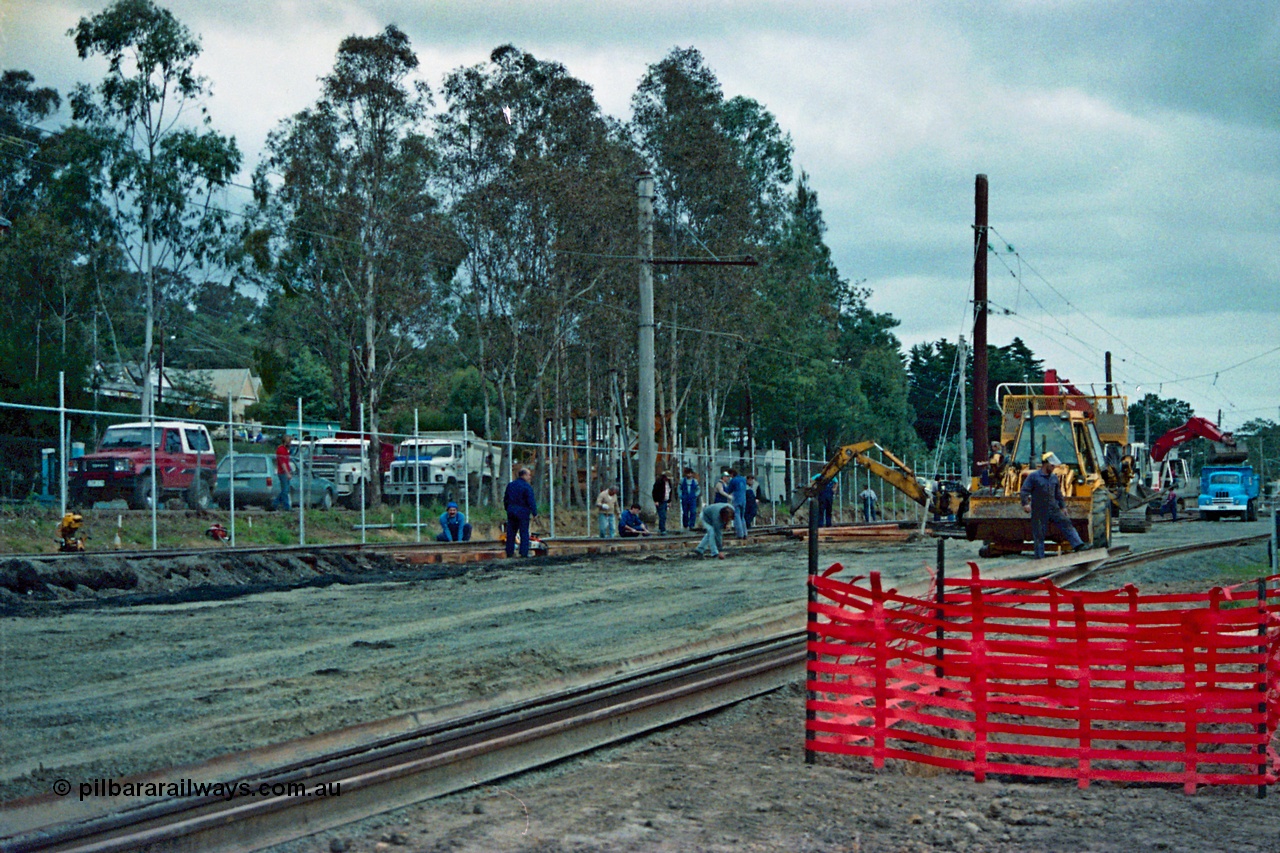 115-01
Hurstbridge, old stabling sidings being removed to make new stabling yard, new fencing, timber traction poles, new sleepers and rail being placed, workers and backhoe, station in the background.
