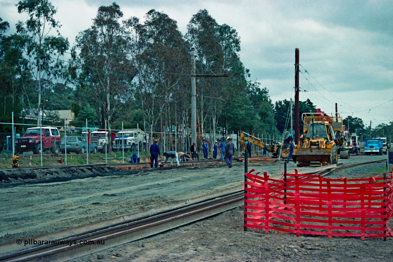 115-02
Hurstbridge, old stabling sidings being removed to make new stabling yard, new fencing, timber traction poles, new sleepers and rail being placed, workers and backhoe, station in the background.
