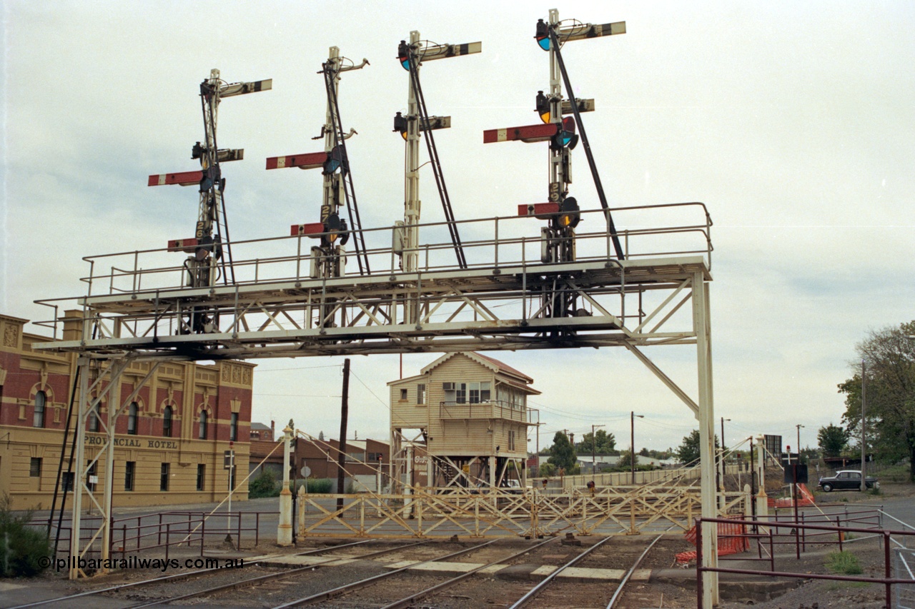 128-04
Ballarat station, view of elevated Ballarat B or Lydiard Street Signal Box, interlocked gates and semaphore signal gantry with semaphore signal Posts 26 and 27 Up signals removed, Post 28 facing away and Post 29, looking west across crib pedestrian crossing, Provincial Hotel on the left.
