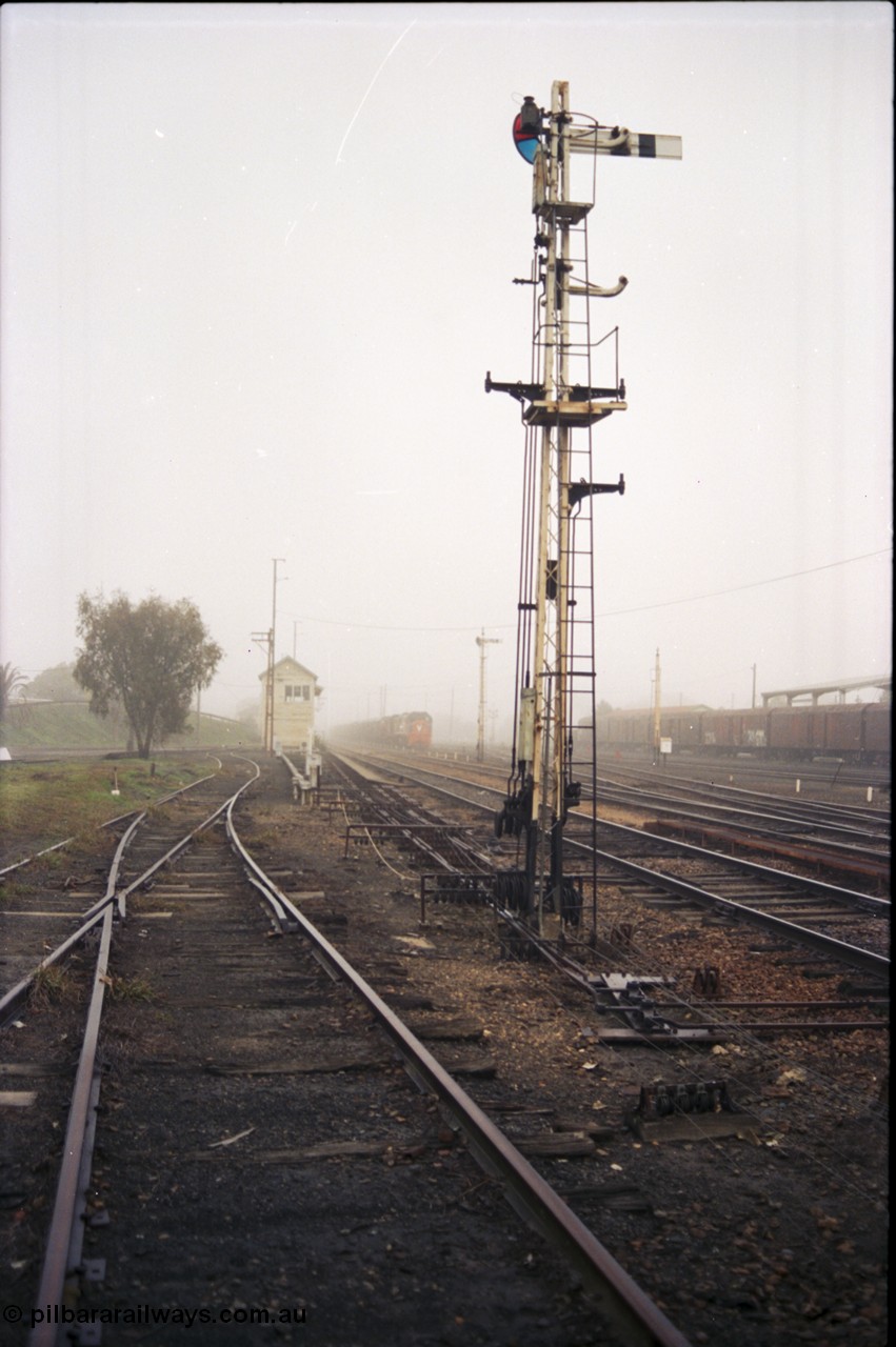 142-1-15
Benalla yard overview, looking beyond a stripped signal post 27, point rodding and wires, tracks to loco depot / workshops at left, Benalla signal box and stabled 9303 goods train in background, very foggy.

