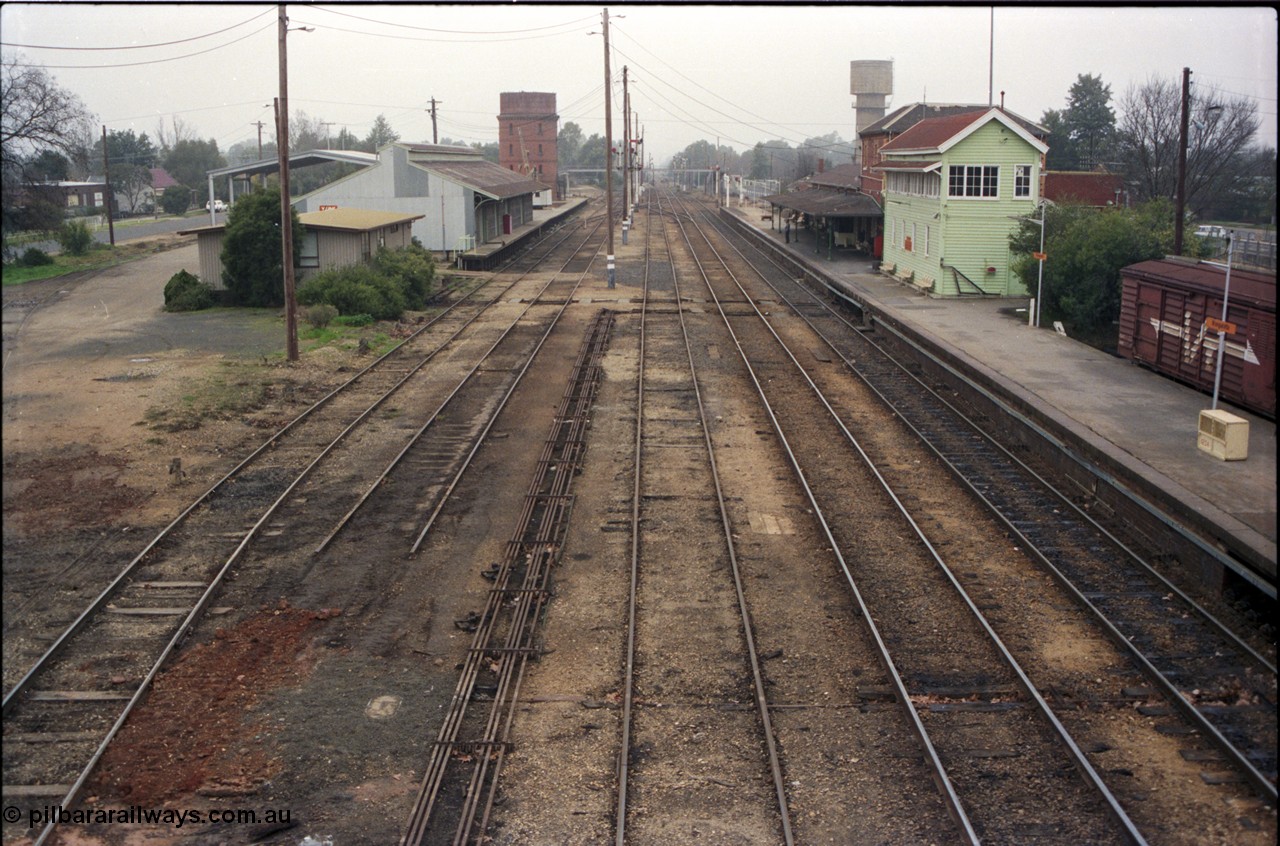 142-2-01
Wangaratta station yard overview looking north from footbridge, No.4 Rd disconnected, goods shed and platform and Freightgate canopy, water tower and crane on the left, station building and elevated signal box on the right, north end footbridge visible in the distance, point rodding running down middle of yard.
