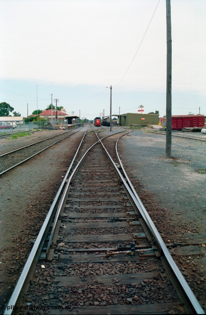 156-08
Shepparton yard view looking south, points for No.5 Road off No.2 Road, then points for No.3 and 4 Roads, engine siding at left, and No.1 Road running down to station platform and building, grounded B van at right, with stabled T class locomotive and fuel waggons with goods shed and Freightgate and cement silos in the background.
