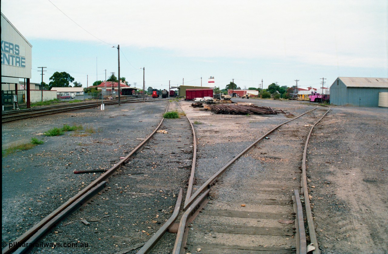 156-10
Shepparton station yard overview looking south down No.5 Road towards the goods shed and stabled T class, fuel train and louvre vans, the former Fruit Siding curves around the back of the goods shed at right, grounded B van and per way stockpiles of rails and pipes in middle of frame, station building and platform on the left, awning of Tubemakers at left over former Engine Track.
