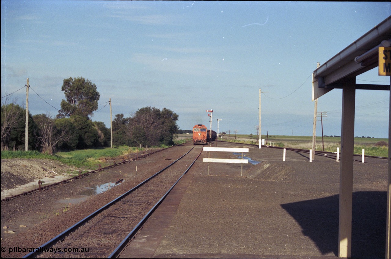 157-03
Gheringhap, view from station platform looking east, the former location of the duplicated line and station platform visible opposite with point rodding and signal wires, V/Line down broad gauge grain train 9121 with a G class leading pulls out of Siding A, semaphore signal posts 3, facing camera and 4, facing away, Sidings B visible on the right running behind station building.
