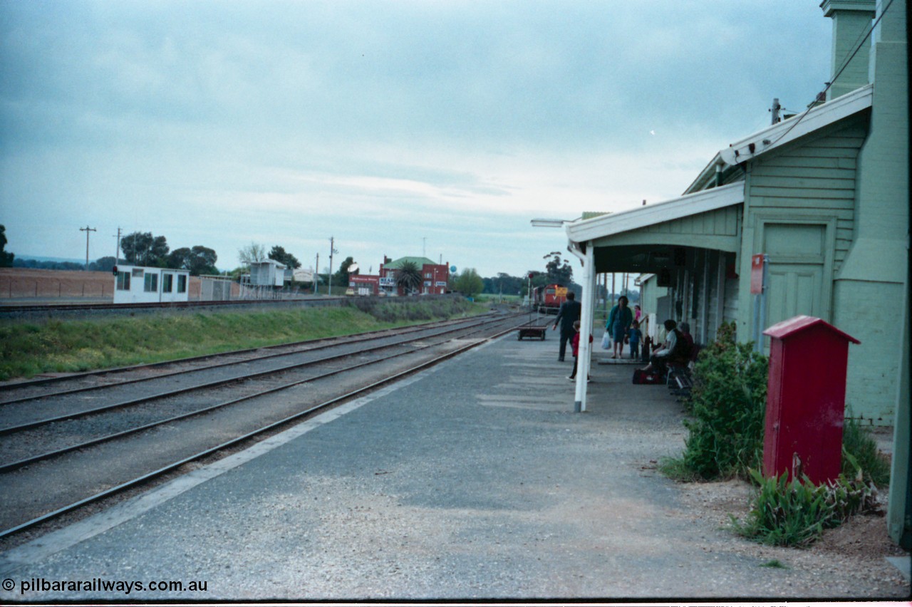 160-07
Murchison East, station platform view of down Cobram passenger train arriving into No.1 Rd, grain receival infrastructure on the left with silos gravitational road, Railway Hotel in the background, passengers awaiting train arrival, station building.
