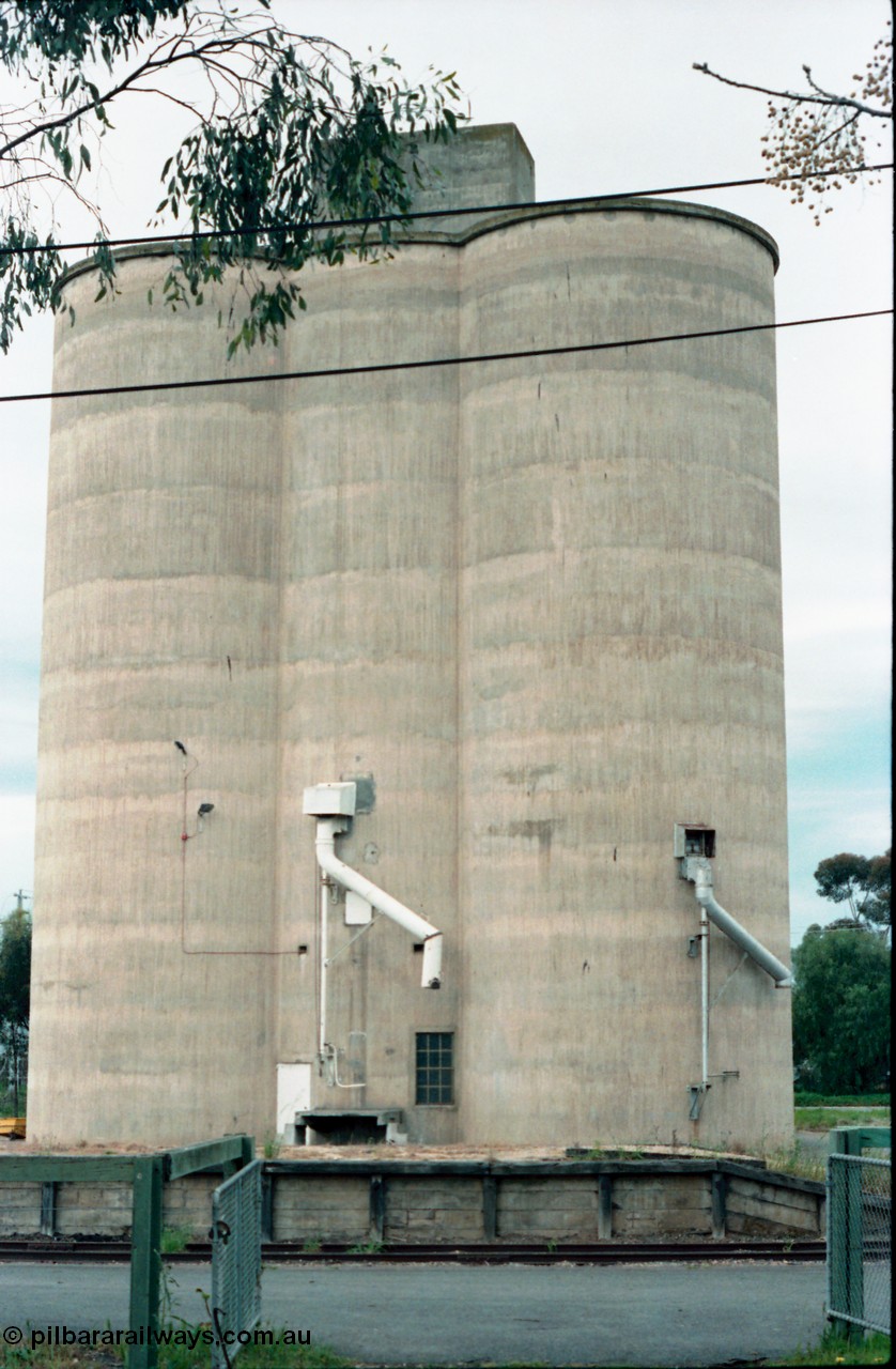 161-03
Numurkah, view from outside platform looking across yard through gates, goods loading platform visible with Williamstown style silo complex located behind it with train loading spouts.

